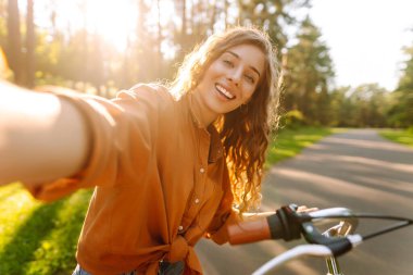 Telefonlu ve bisikletli neşeli kadın güneşli parkta selfie çekiyor. Bisiklete binen ve dışarıda havanın tadını çıkaran genç bir kadın. Yürüme konsepti, selfie. Etkin yaşam biçimi.