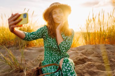 Şapkalı neşeli kadın telefonda kumlu bir kumsalın arka planında selfie çekiyor. Güzel bir kadın günbatımında setin üzerinde yürüyor. Bloglama ve boş zaman kavramı.