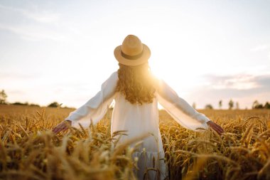 Beyaz elbiseli ve hasır şapkalı bir kadının altın buğday tarlasında dikiz aynasından görünüşü. Güzel bir kadın gün batımından zevk alır ve açık havada özgür hisseder. Güzellik kavramı, özgürlük, takip et.