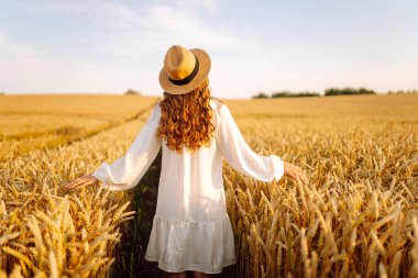 Beyaz elbiseli ve hasır şapkalı bir kadının altın buğday tarlasında dikiz aynasından görünüşü. Güzel bir kadın gün batımından zevk alır ve açık havada özgür hisseder. Güzellik kavramı, özgürlük, takip et.