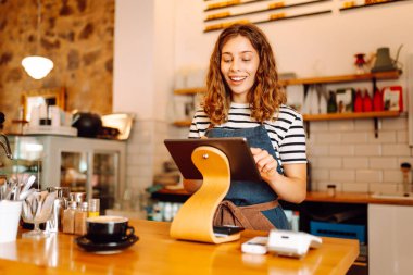 Genç bir bayan barmen hazır kahveyle bir barın arkasında duruyor. Dijital tabletli güzel bir kadın sıcak bir kahve dükkanında güneş ışığının tadını çıkarıyor. Küçük işletme, içecek konsepti.
