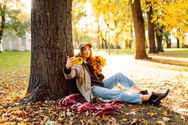 Yağmurluk giyen, telefonlu genç bir kadın sonbahar parkında bir ağacın yanındaki battaniyenin üzerinde oturuyor. Kadın selfie ya da blog çekiyor ve açık havada yürümekten hoşlanıyor. Sonbahar konsepti, teknoloji.