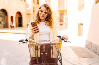 Parkta güneşli bir günde elinde telefon olan neşeli bir turist bisiklete biniyor. Genç bir kadın gün batımında yürümekten, bloglardan ya da telefonundaki mesajlardan hoşlanır. Blogculuk, turizm konsepti.