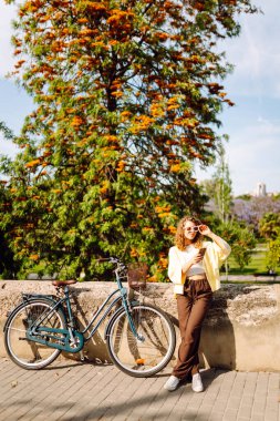 Parkta güneşli bir günde elinde telefon olan neşeli bir turist bisiklete biniyor. Genç bir kadın gün batımında yürümekten, bloglardan ya da telefonundaki mesajlardan hoşlanır. Blogculuk, turizm konsepti.