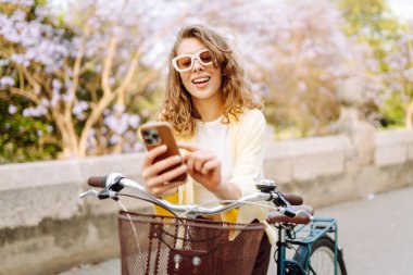 Parkta güneşli bir günde elinde telefon olan neşeli bir turist bisiklete biniyor. Genç bir kadın gün batımında yürümekten, bloglardan ya da telefonundaki mesajlardan hoşlanır. Blogculuk, turizm konsepti.