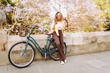 Parkta güneşli bir günde elinde telefon olan neşeli bir turist bisiklete biniyor. Genç bir kadın gün batımında yürümekten, bloglardan ya da telefonundaki mesajlardan hoşlanır. Blogculuk, turizm konsepti.