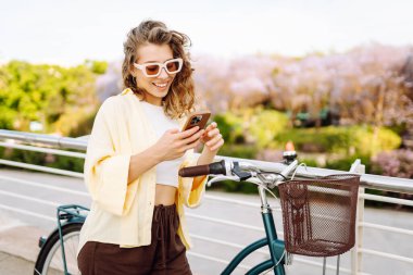 Parkta güneşli bir günde elinde telefon olan neşeli bir turist bisiklete biniyor. Genç bir kadın gün batımında yürümekten, bloglardan ya da telefonundaki mesajlardan hoşlanır. Blogculuk, turizm konsepti.