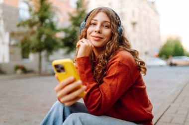 Güzel bir kadının portresi. Kablosuz kulaklıkları ve açık havada yürüyüş yapan bir telefonu var. Şehrin sokaklarında eğlenen ve müzik dinleyen bir turist. Teknoloji ve yürüme kavramı.