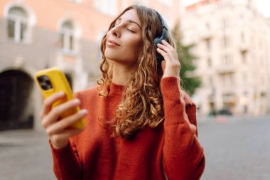Güzel bir kadının portresi. Kablosuz kulaklıkları ve açık havada yürüyüş yapan bir telefonu var. Şehrin sokaklarında eğlenen ve müzik dinleyen bir turist. Teknoloji ve yürüme kavramı.