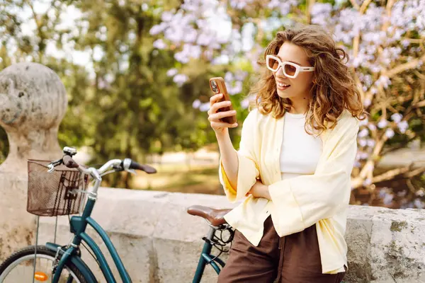 Parkta güneşli bir günde elinde telefon olan neşeli bir turist bisiklete biniyor. Genç bir kadın gün batımında yürümekten, bloglardan ya da telefonundaki mesajlardan hoşlanır. Blogculuk, turizm konsepti.