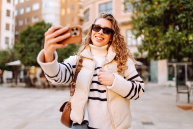 Telefonu olan neşeli bir turist açık havada güneşli bir sokakta yürüyor. Genç bir kadın telefonuyla blog yazıyor ve günbatımının tadını çıkarıyor. Blogculuk, turizm ve hafta sonu konsepti.