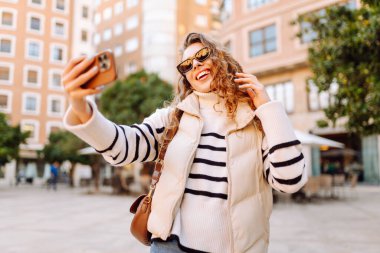 Telefonu olan neşeli bir turist açık havada güneşli bir sokakta yürüyor. Genç bir kadın telefonuyla blog yazıyor ve günbatımının tadını çıkarıyor. Blogculuk, turizm ve hafta sonu konsepti.