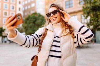 Telefonu olan neşeli bir turist açık havada güneşli bir sokakta yürüyor. Genç bir kadın telefonuyla blog yazıyor ve günbatımının tadını çıkarıyor. Blogculuk, turizm ve hafta sonu konsepti.