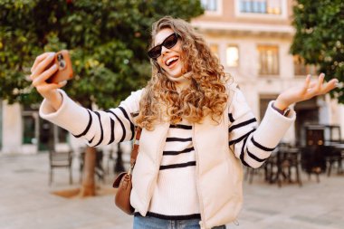 Telefonu olan neşeli bir turist açık havada güneşli bir sokakta yürüyor. Genç bir kadın telefonuyla blog yazıyor ve günbatımının tadını çıkarıyor. Blogculuk, turizm ve hafta sonu konsepti.