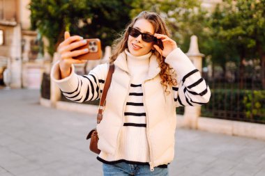 Telefonu olan neşeli bir turist açık havada güneşli bir sokakta yürüyor. Genç bir kadın telefonuyla blog yazıyor ve günbatımının tadını çıkarıyor. Blogculuk, turizm ve hafta sonu konsepti.