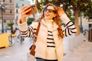 Telefonu olan neşeli bir turist açık havada güneşli bir sokakta yürüyor. Genç bir kadın telefonuyla blog yazıyor ve günbatımının tadını çıkarıyor. Blogculuk, turizm ve hafta sonu konsepti.