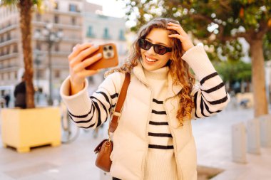 Telefonu olan neşeli bir turist açık havada güneşli bir sokakta yürüyor. Genç bir kadın telefonuyla blog yazıyor ve günbatımının tadını çıkarıyor. Blogculuk, turizm ve hafta sonu konsepti.