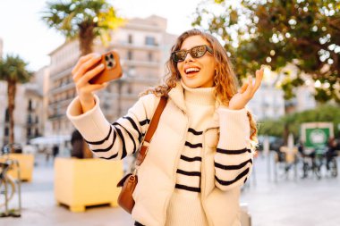 Telefonu olan neşeli bir turist açık havada güneşli bir sokakta yürüyor. Genç bir kadın telefonuyla blog yazıyor ve günbatımının tadını çıkarıyor. Blogculuk, turizm ve hafta sonu konsepti.