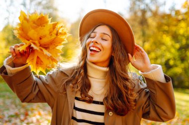 Şapkalı şık bir kadın ve elinde bir buket yaprakla sonbahar parkında günbatımının tadını çıkarıyor. Güzel bir kadın eğleniyor ve dışarı çıkıyor. Mevsim kavramı. Yaşam biçimi.