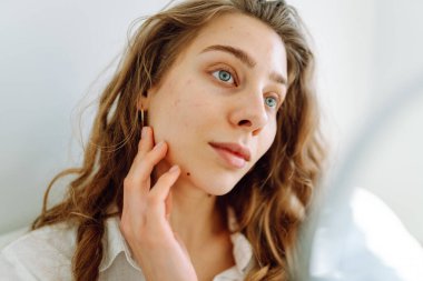 Samimi bir odada küçük bir aynaya bakan genç bir kadının portresi. Güzel bir kadın elleriyle yüzüne dokunur ve doğal güzellikten zevk alır. Güzellik kavramı, dermatoloji, rahatlık.