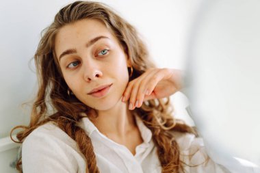 Samimi bir odada küçük bir aynaya bakan genç bir kadının portresi. Güzel bir kadın elleriyle yüzüne dokunur ve doğal güzellikten zevk alır. Güzellik kavramı, dermatoloji, rahatlık.