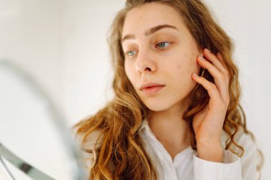 Samimi bir odada küçük bir aynaya bakan genç bir kadının portresi. Güzel bir kadın elleriyle yüzüne dokunur ve doğal güzellikten zevk alır. Güzellik kavramı, dermatoloji, rahatlık.