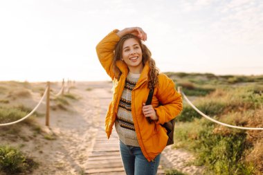 Sırt çantalı kadın yürüyüşçü deniz manzarasına karşı yürüyen ahşap yolda. Mutlu kadın gün batımında yürüyor ve maceranın tadını çıkarıyor. Yürüyüş konsepti. Etkin yaşam biçimi.