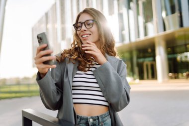 Gözlüklü ve telefonlu bir kadın modern bir binanın arka planında merdivenlerde oturuyor. Neşeli kadın akıllı telefon kullanıyor, blog yazıyor ve güneşli bir günün tadını çıkarıyor. İş konsepti.