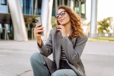 Gözlüklü ve telefonlu bir kadın modern bir binanın arka planında merdivenlerde oturuyor. Neşeli kadın akıllı telefon kullanıyor, blog yazıyor ve güneşli bir günün tadını çıkarıyor. İş konsepti.