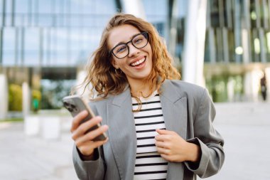 Gözlüklü ve telefonlu bir kadın modern bir binanın arka planında merdivenlerde oturuyor. Neşeli kadın akıllı telefon kullanıyor, blog yazıyor ve güneşli bir günün tadını çıkarıyor. İş konsepti.