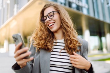 Gözlüklü ve telefonlu bir kadın modern bir binanın arka planında merdivenlerde oturuyor. Neşeli kadın akıllı telefon kullanıyor, blog yazıyor ve güneşli bir günün tadını çıkarıyor. İş konsepti.