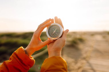 Gün batımındaki bir plajın arka planına pusula tutan genç bir kadın yolcunun ellerine yakın çekim. Parlak ceketli bir kadın seyahat yönüne bakar ve güneş ışığından hoşlanır. Seyahat kavramı.