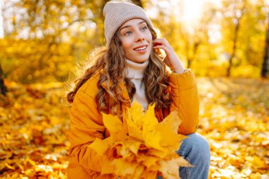 Parlak ceketli neşeli bir kadın güneşli bir parkta toplanır ve parlak yapraklar saçar. Güzel bir kadın eğleniyor ve sonbahar manzarasının tadını çıkarıyor. Yürüyüş, seyahat ve rahatlama kavramı..