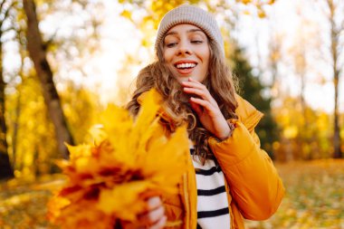 Parlak ceketli neşeli bir kadın güneşli bir parkta toplanır ve parlak yapraklar saçar. Güzel bir kadın eğleniyor ve sonbahar manzarasının tadını çıkarıyor. Yürüyüş, seyahat ve rahatlama kavramı..