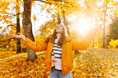 Parlak ceketli neşeli bir kadın güneşli bir parkta toplanır ve parlak yapraklar saçar. Güzel bir kadın eğleniyor ve sonbahar manzarasının tadını çıkarıyor. Yürüyüş, seyahat ve rahatlama kavramı..