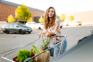 Gün batımında bagajına taze sebze ve meyve koyan tatlı bir kadın. Ellerinde ananas, otoparkta bir çanta dolusu ürünü olan mutlu bir ev hanımı. Alışveriş, rutin konsept.
