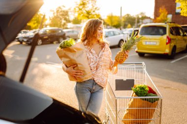 Gün batımında bagajına taze sebze ve meyve koyan tatlı bir kadın. Ellerinde ananas, otoparkta bir çanta dolusu ürünü olan mutlu bir ev hanımı. Alışveriş, rutin konsept.