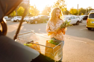 Gün batımında bagajına taze sebze ve meyve koyan tatlı bir kadın. Ellerinde ananas, otoparkta bir çanta dolusu ürünü olan mutlu bir ev hanımı. Alışveriş, rutin konsept.
