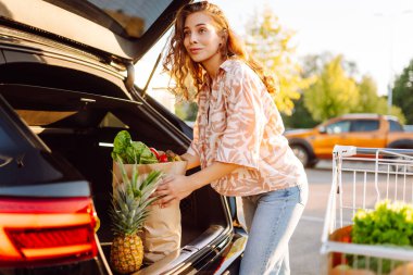 Gün batımında bagajına taze sebze ve meyve koyan tatlı bir kadın. Ellerinde ananas, otoparkta bir çanta dolusu ürünü olan mutlu bir ev hanımı. Alışveriş, rutin konsept.