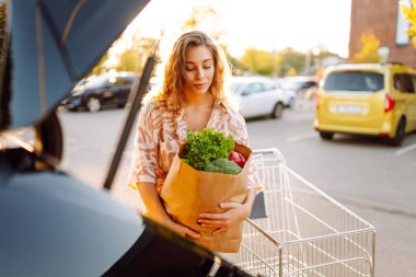 Gün batımında bagajına taze sebze ve meyve koyan tatlı bir kadın. Ellerinde ananas, otoparkta bir çanta dolusu ürünü olan mutlu bir ev hanımı. Alışveriş, rutin konsept.