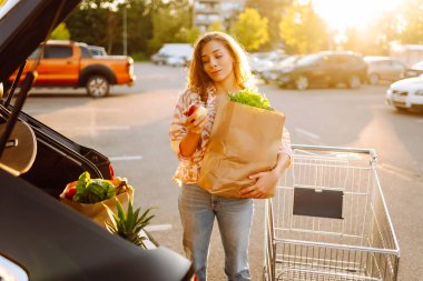 Gün batımında bagajına taze sebze ve meyve koyan tatlı bir kadın. Ellerinde ananas, otoparkta bir çanta dolusu ürünü olan mutlu bir ev hanımı. Alışveriş, rutin konsept.