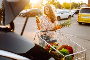 Gün batımında bagajına taze sebze ve meyve koyan tatlı bir kadın. Ellerinde ananas, otoparkta bir çanta dolusu ürünü olan mutlu bir ev hanımı. Alışveriş, rutin konsept.