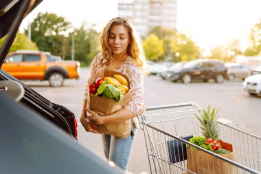 Gün batımında bagajına taze sebze ve meyve koyan tatlı bir kadın. Ellerinde ananas, otoparkta bir çanta dolusu ürünü olan mutlu bir ev hanımı. Alışveriş, rutin konsept.