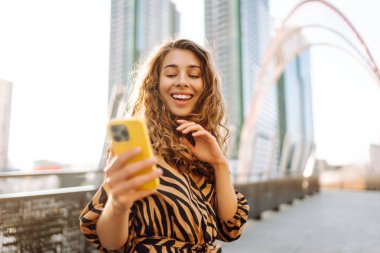 Gün batımında köprüde telefonu olan neşeli bir kadın. Genç turist akıllı telefondan blog yazıyor ve havanın tadını çıkarıyor. Blogculuk, turizm ve teknoloji kavramı.