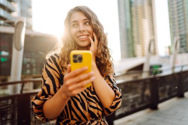 Gün batımında köprüde telefonu olan neşeli bir kadın. Genç turist akıllı telefondan blog yazıyor ve havanın tadını çıkarıyor. Blogculuk, turizm ve teknoloji kavramı.