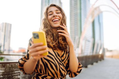 Gün batımında köprüde telefonu olan neşeli bir kadın. Genç turist akıllı telefondan blog yazıyor ve havanın tadını çıkarıyor. Blogculuk, turizm ve teknoloji kavramı.