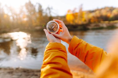 Genç bayan yürüyüşçünün ellerinin gölün arka planına pusula tutarken yakın plan görüntüleri. Parlak ceketli kadın günbatımının tadını çıkarıyor ve güneşli sonbahar patikasında yürüyüş yönü arıyor. Navigasyon kavramı.