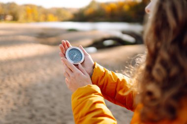 Genç bayan yürüyüşçünün ellerinin gölün arka planına pusula tutarken yakın plan görüntüleri. Parlak ceketli kadın günbatımının tadını çıkarıyor ve güneşli sonbahar patikasında yürüyüş yönü arıyor. Navigasyon kavramı.