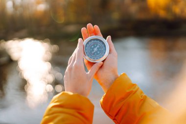 Genç bayan yürüyüşçünün ellerinin gölün arka planına pusula tutarken yakın plan görüntüleri. Parlak ceketli kadın günbatımının tadını çıkarıyor ve güneşli sonbahar patikasında yürüyüş yönü arıyor. Navigasyon kavramı.