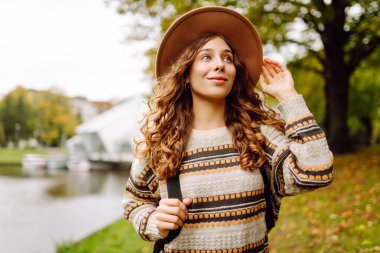 Kıvırcık saçlı ve şapkalı mutlu kadın parkta sonbahar gününün tadını çıkarıyor. Göl kenarındaki bir çayırda yürüyen sırt çantalı güzel bir turist. Etrafı yapraklarla çevrili. Yürüyüş kavramı, turizm.